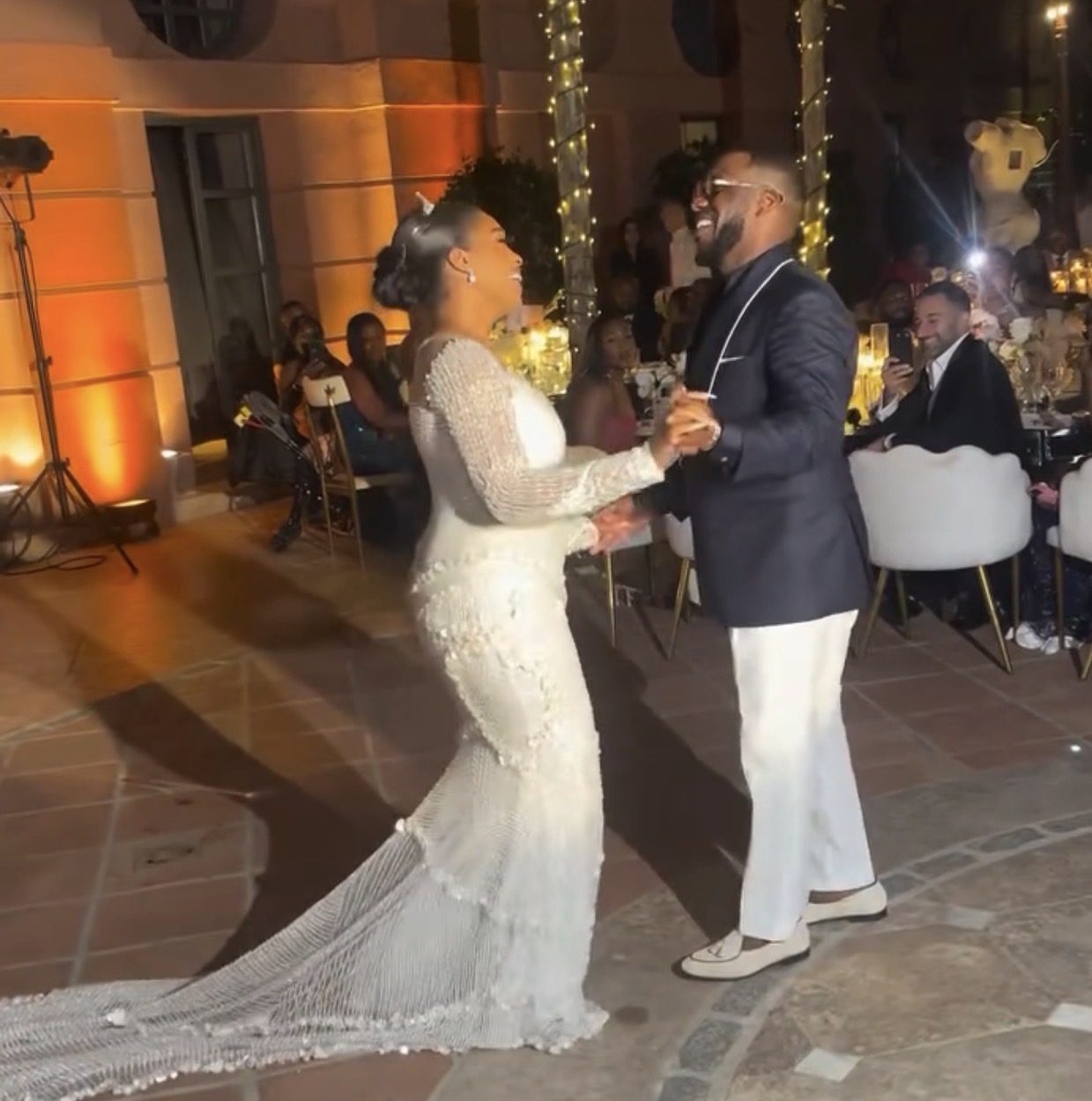 A father and daughter dancing joyfully on a sunlit terrace in Spain, celebrating a wedding with warm lighting and floral decorations.