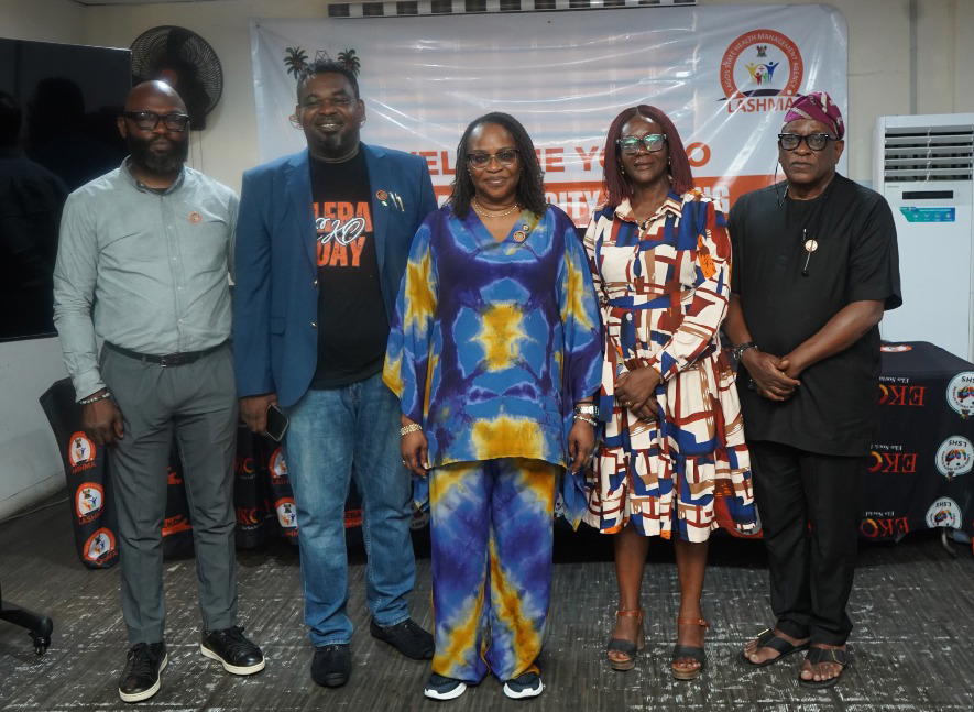 A diverse group of journalists and healthcare workers discussing ILERA EKO health insurance in a Lagos community setting, with a modern hospital and health awareness billboard in the background.