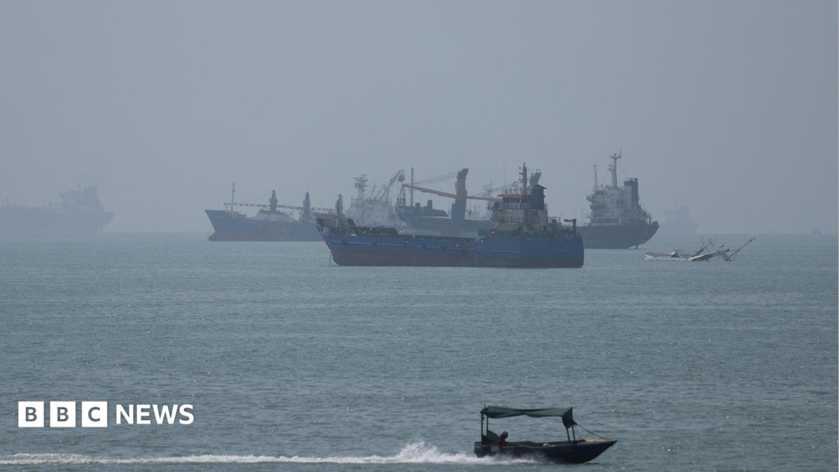 Aerial view of an oil tanker in the Strait of Hormuz with naval ships in the background and a distant explosion on Larak Island.