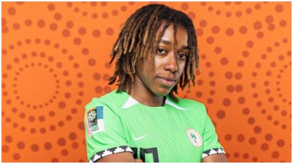 A Nigerian flag waves in the foreground as a blurred footballer in a PSG kit dribbles a ball on a brightly lit pitch, with Manchester City’s crest visible in the background.
