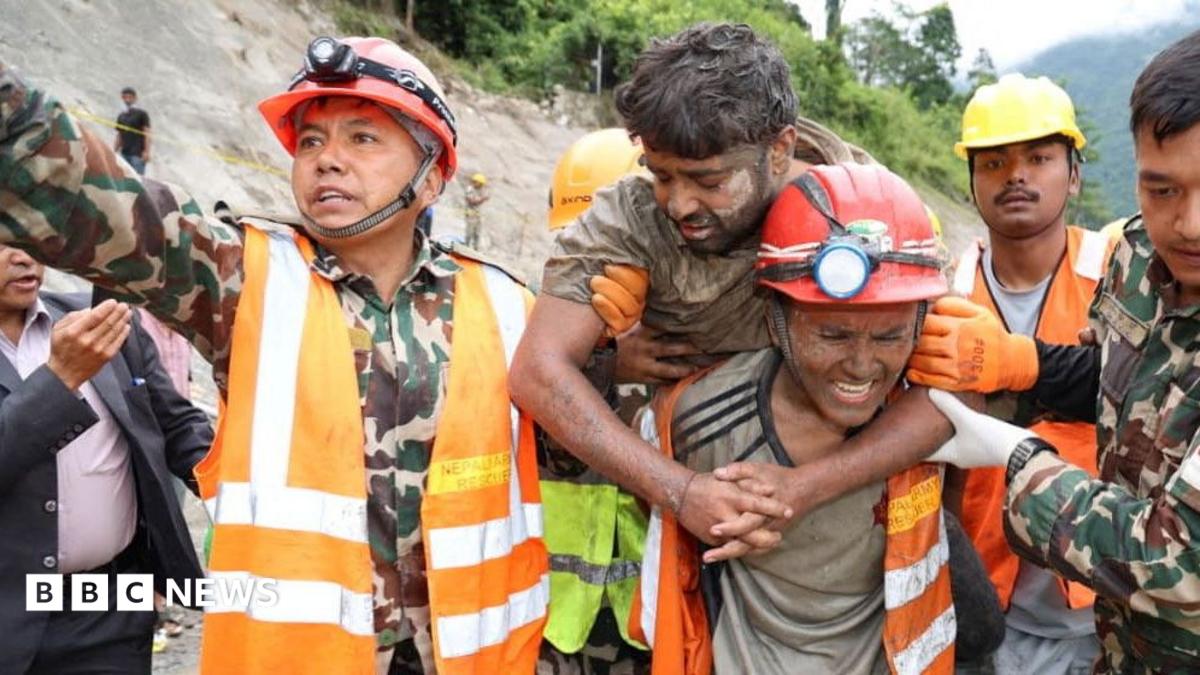 Rescue workers guide flood survivors out of a riverside tunnel entrance in Nepal, ropes and helmets visible on a muddy slope.