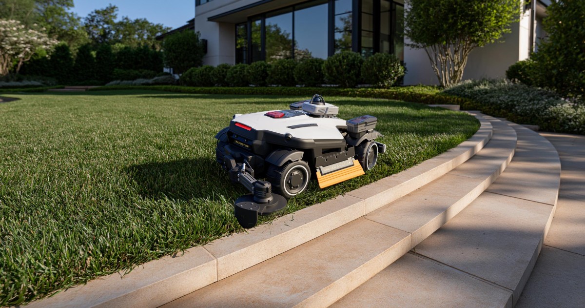 A black robot lawnmower with an extendable edge-trimming arm mowing along a flowerbed next to a paved driveway, showcasing its precision trimming capability.