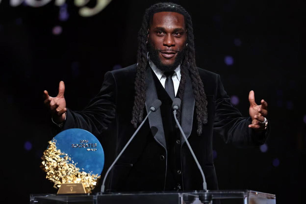 Stage at the 2026 Ballon d’Or ceremony with a trophy and microphone, symbolising Burna Boy presenting the Socrates Award to Bukayo Saka.