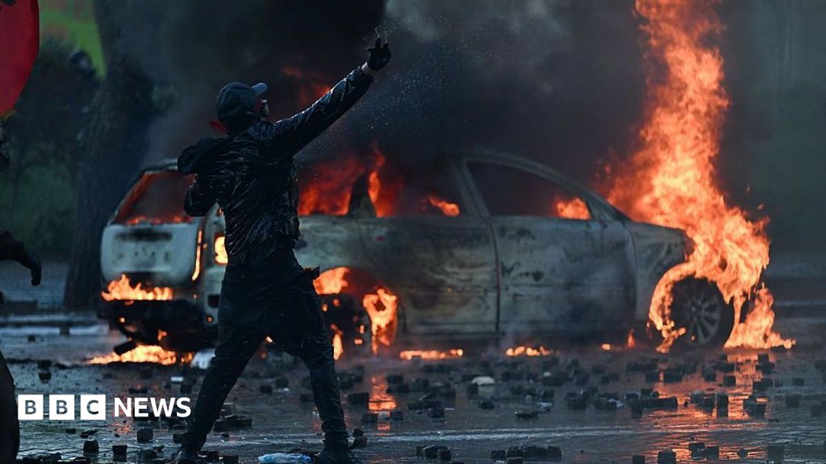 Nighttime street in Tirana with police lights and protest signs after violent clashes.